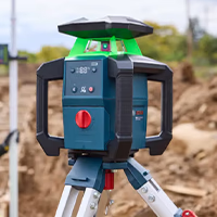 A blue and black Bosch rotary laser level with a bright green laser emitter housing, mounted on a silver aluminum tripod at an outdoor construction site.